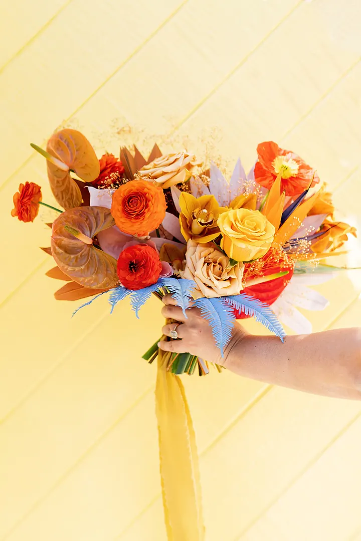 A hand holding a bouquet of orange, red, and gold flowers with pale blue fern leaves against a yellow wall.
