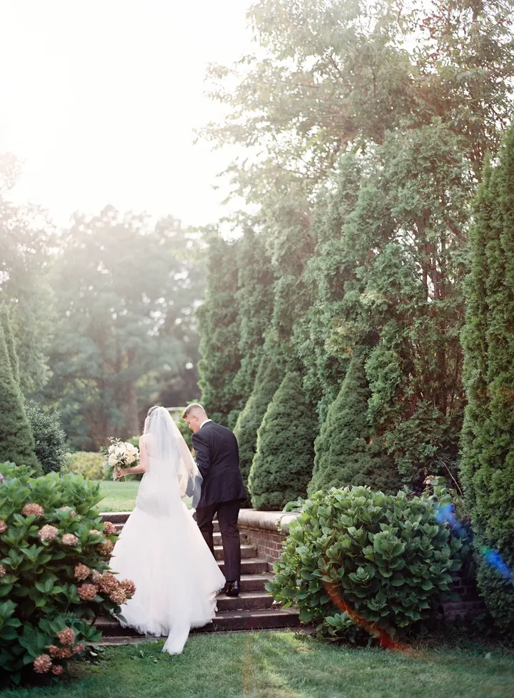 A bride and groom walking up garden steps past hydrangeas and tall green hedges.