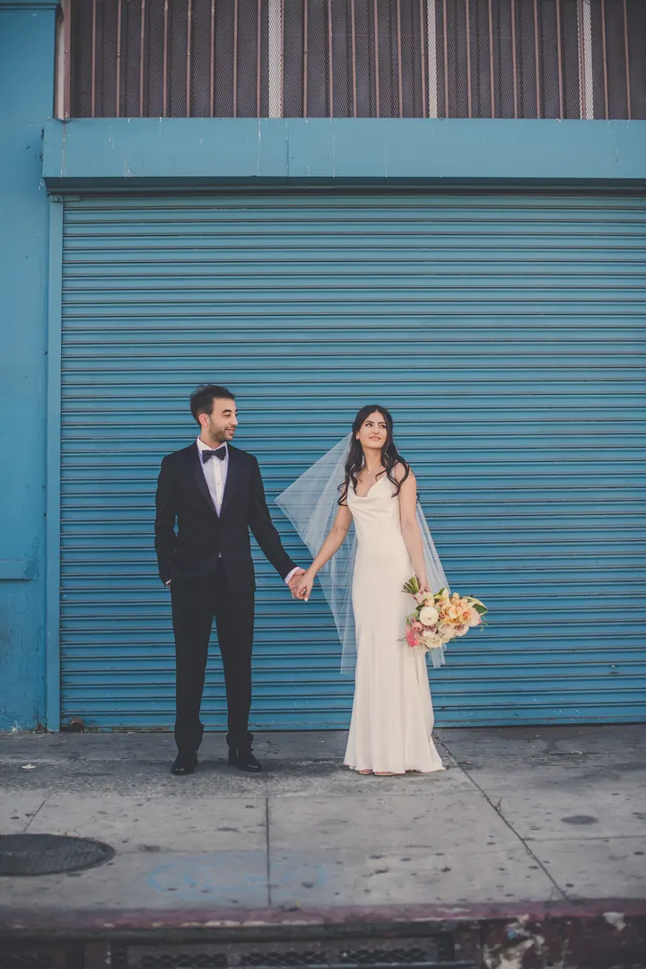 A couple holding hands in front of a teal roll-up door, the bride's veil lifting in the breeze.