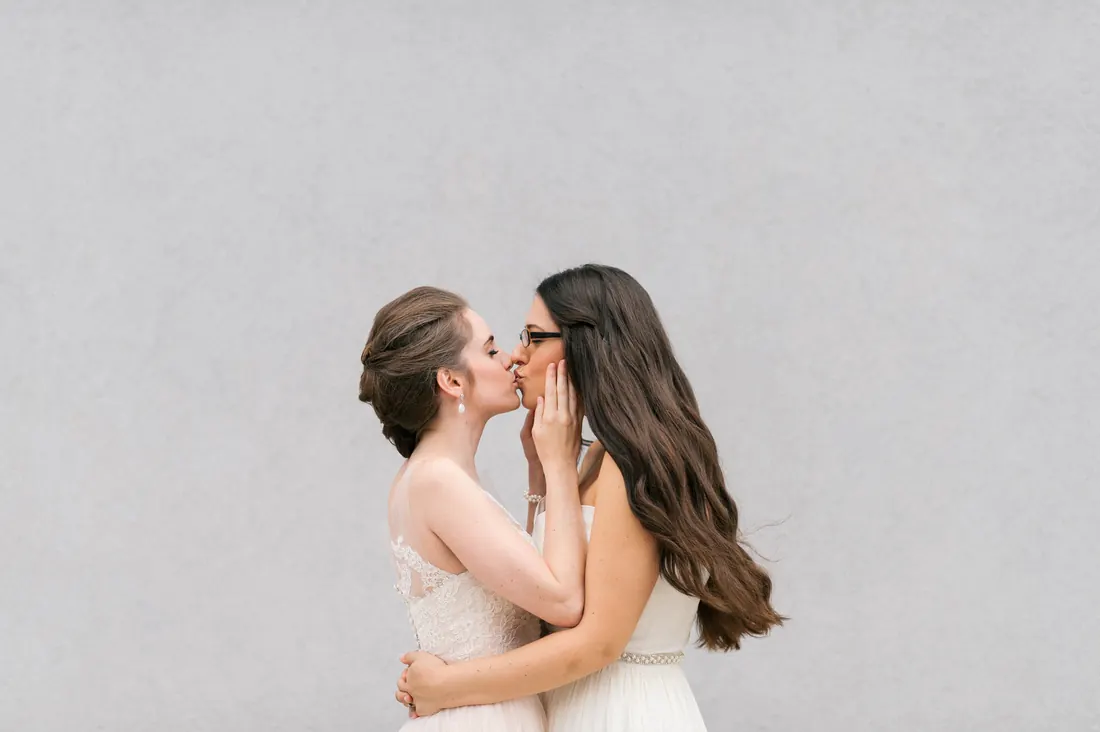 Two brides kissing against a soft gray wall.