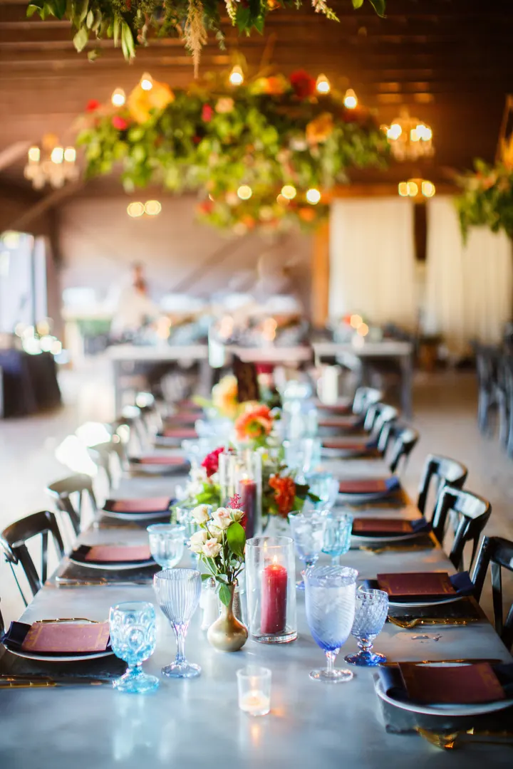 A long reception table set with blue glassware, red candles, and a hanging installation of flowers and string lights.