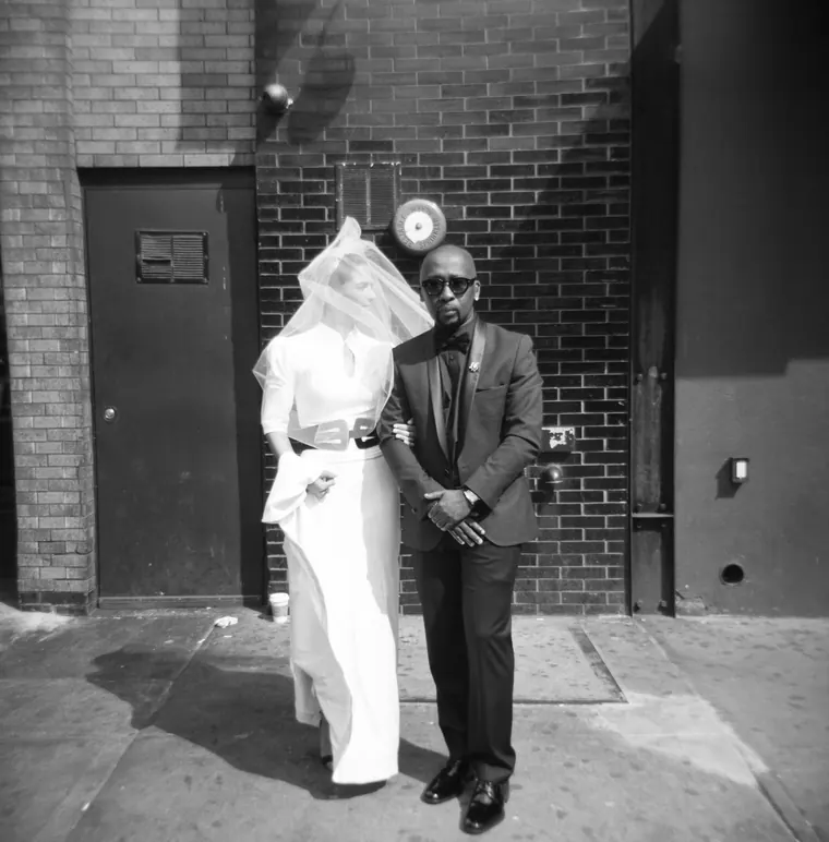 A black-and-white portrait of a bride in a veil arm in arm with a groom in sunglasses and a tuxedo, in front of a brick wall.