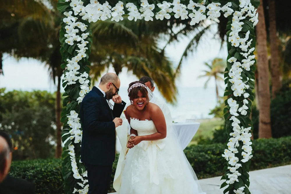 A bride laughs while a groom wipes a tear under an arch covered in white orchids.