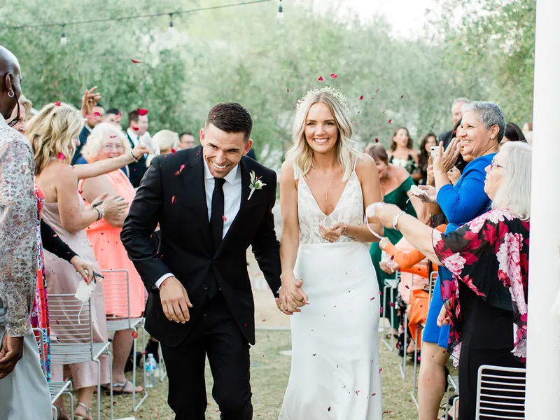 A bride and groom walk down an outdoor aisle holding hands as guests toss petals.